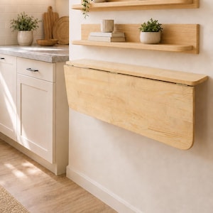 May include: A light-colored wooden wall-mounted drop-leaf table with rounded edges, positioned below two matching wooden floating shelves. The shelves hold small potted green plants, a ceramic vase, a small bowl, and two stacked books.