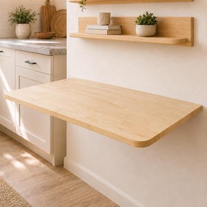 May include: A light-colored wooden wall-mounted drop-leaf table attached to a wall below two matching wooden floating shelves. The shelves hold small potted plants, a ceramic vase, a small bowl, and a stack of two books with a ribbed cup on top.