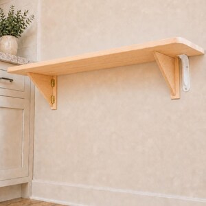 May include: A light-colored wooden wall-mounted folding shelf featuring a smooth rectangular surface, supported by two triangular wooden brackets. The left bracket is attached with gold-toned metal hinges, while the right bracket is secured with a white metal support arm, allowing the shelf to be folded down against the wall.