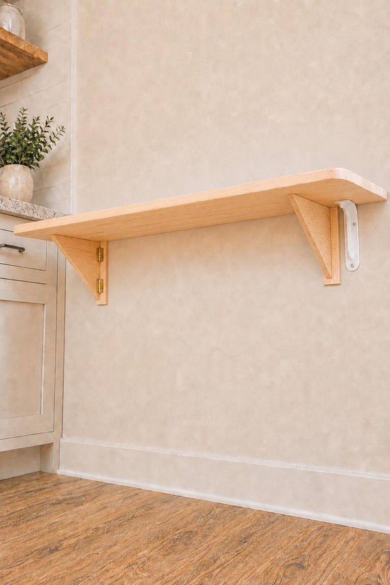 May include: A light-colored wooden wall-mounted folding shelf featuring a smooth rectangular surface, supported by two triangular wooden brackets. The left bracket is attached with gold-toned metal hinges, while the right bracket is secured with a white metal support arm, allowing the shelf to be folded down against the wall.