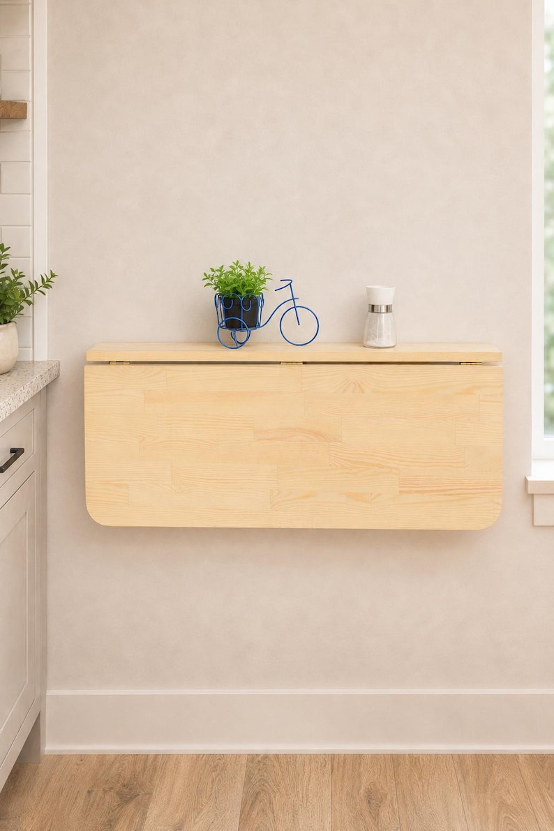 May include: A light-colored wooden wall-mounted drop-leaf table with rounded corners. The table is folded down against the wall. On top of the table sits a blue wire bicycle-shaped plant holder with a green plant and a clear glass salt shaker.