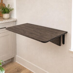 May include: A wall-mounted, rectangular, dark wood-toned folding table featuring rounded corners and a smooth surface. The table is attached to the wall with black metal support brackets, allowing it to be positioned horizontally for use.