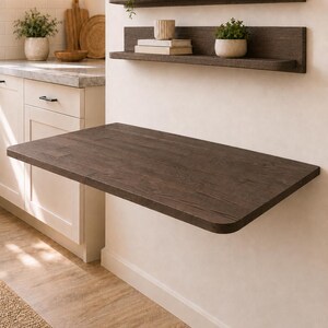 May include: A dark brown wood-grain wall-mounted floating desk with rounded corners, positioned below two matching horizontal wall shelves. The shelves hold small potted green plants, a white ceramic vase, a bowl, and two stacked books.