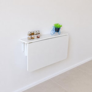 May include: A white wall-mounted folding table with a smooth rectangular surface. Four small glass spice jars with labels are arranged in a row on the top shelf, next to a decorative blue wire bicycle sculpture holding a small green plant.