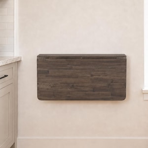 May include: A rectangular, dark brown, wood-grain wall-mounted folding table. The table is in a closed, vertical position against a light-colored wall, featuring a smooth, flat surface with visible wood grain patterns and rounded corners.