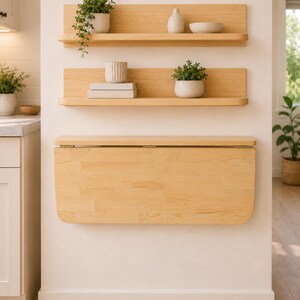 May include: Two light-colored wooden wall shelves with rounded edges and a matching wall-mounted folding table positioned below. The shelves hold small potted plants, a white vase, a white bowl, and two stacked books with white covers.