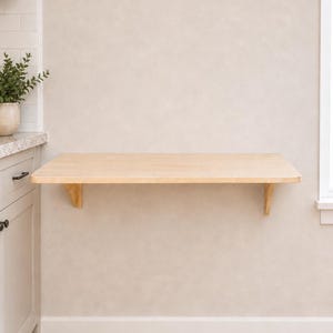 May include: A wall-mounted floating desk featuring a rectangular light-toned wood surface supported by two matching wooden brackets. The desk is installed horizontally against a plain wall, providing a flat, open workspace area for various uses.