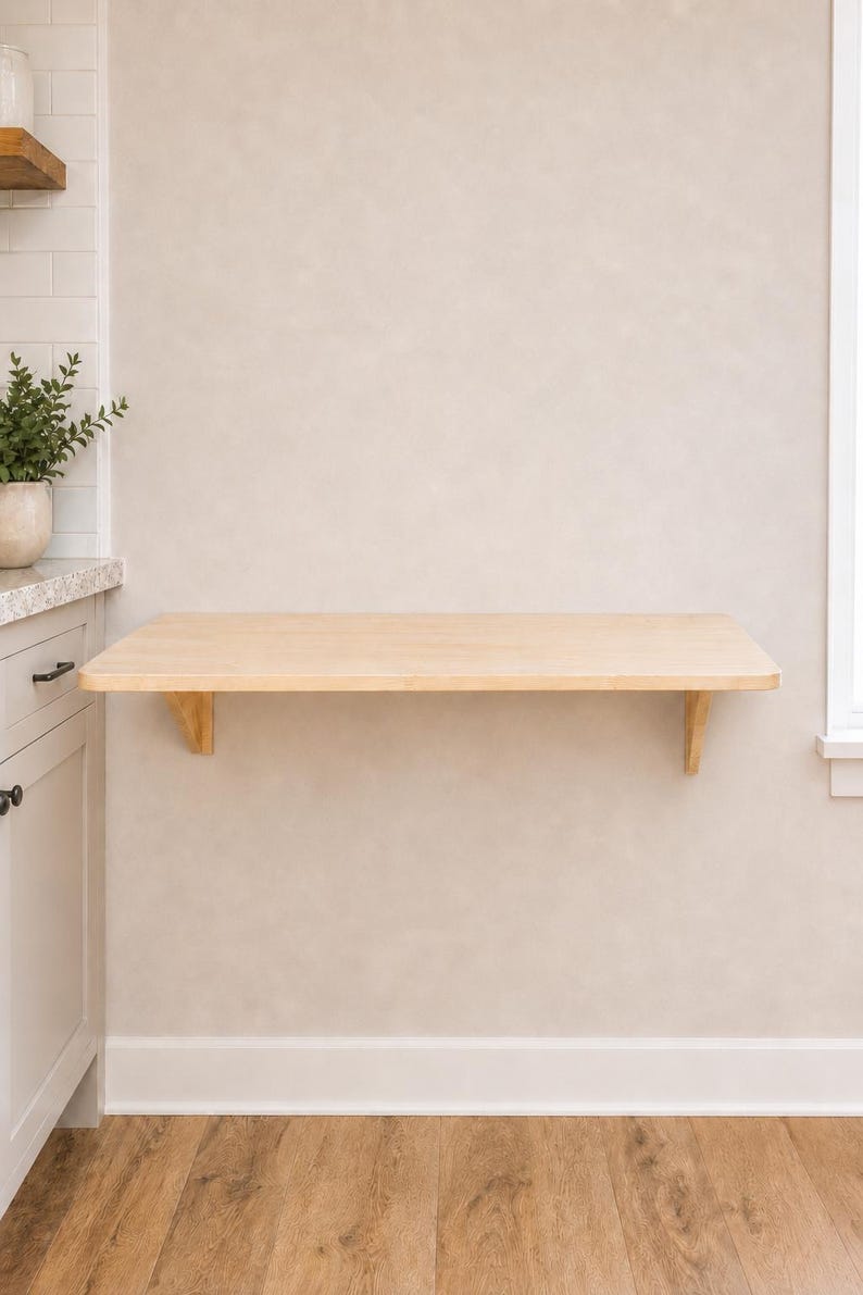 May include: A wall-mounted floating desk featuring a rectangular light-toned wood surface supported by two matching wooden brackets. The desk is installed horizontally against a plain wall, providing a flat, open workspace area for various uses.