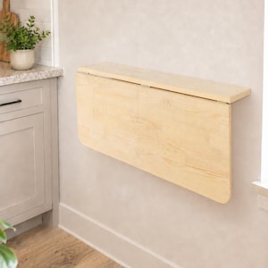 May include: A light-colored wooden wall-mounted drop-leaf table featuring a rectangular top and a hinged panel that folds downward. The table is constructed from natural wood with visible grain patterns and is attached to a flat vertical surface.