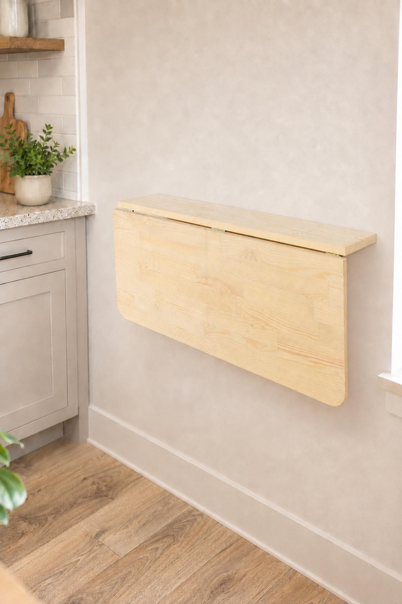 May include: A light-colored wooden wall-mounted drop-leaf table featuring a rectangular top and a hinged panel that folds downward. The table is constructed from natural wood with visible grain patterns and is attached to a flat vertical surface.