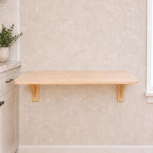 May include: A light-colored wooden wall-mounted floating desk featuring a rectangular surface supported by two matching wooden brackets attached to the wall. The desk has a smooth, natural wood grain finish and a clean, simple rectangular design.
