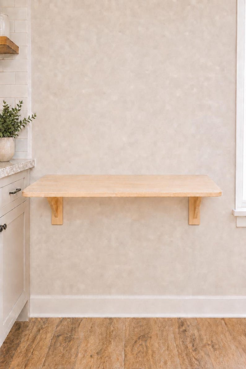 May include: A light-colored wooden wall-mounted floating desk featuring a rectangular surface supported by two matching wooden brackets attached to the wall. The desk has a smooth, natural wood grain finish and a clean, simple rectangular design.