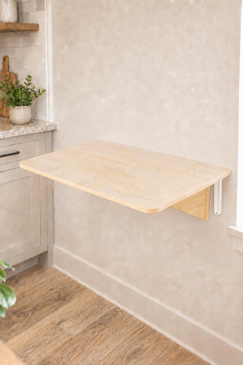 May include: A wall-mounted, rectangular, light-colored wooden folding table with rounded corners. The table is attached to the wall with white metal brackets, featuring a smooth, natural wood grain finish and a flat, horizontal surface.