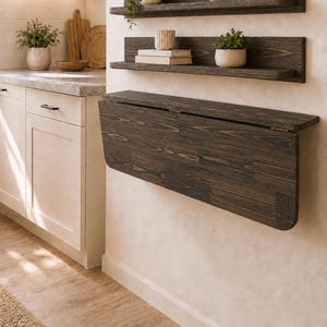 May include: Two dark brown wooden wall-mounted shelves and one dark brown wooden drop-leaf table mounted to a wall. The shelves hold small potted plants, a ceramic vase, a bowl, and two books. The table is in a folded, downward position.