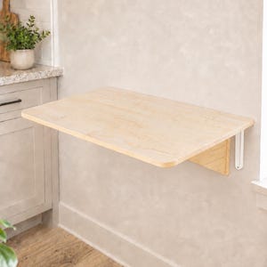 May include: A wall-mounted, rectangular, light-colored wooden folding table with rounded corners. The table is attached to the wall with white metal brackets, featuring a smooth, natural wood grain finish and a flat, horizontal surface.