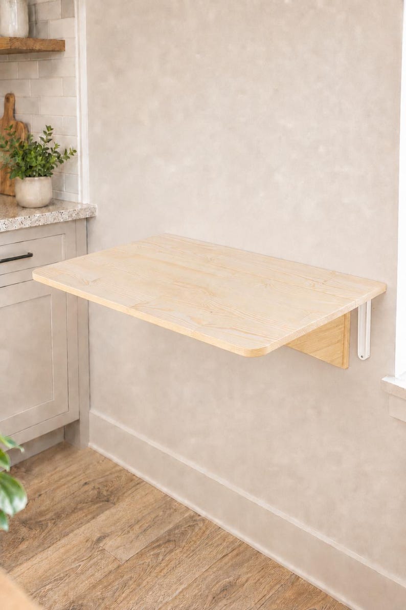 May include: A wall-mounted, rectangular, light-colored wooden folding table with rounded corners. The table is attached to the wall with white metal brackets, featuring a smooth, natural wood grain finish and a flat, horizontal surface.