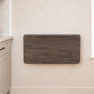 May include: A rectangular, dark brown wooden wall-mounted table in a folded, vertical position. The table features a visible wood grain pattern and rounded corners on the bottom edge, mounted flush against a plain, light-colored interior wall.