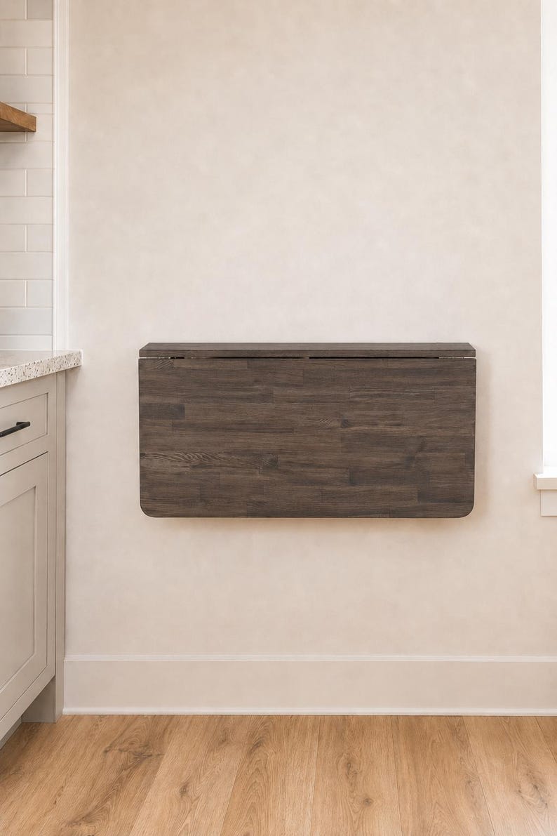 May include: A rectangular, dark brown wooden wall-mounted table in a folded, vertical position. The table features a visible wood grain pattern and rounded corners on the bottom edge, mounted flush against a plain, light-colored interior wall.
