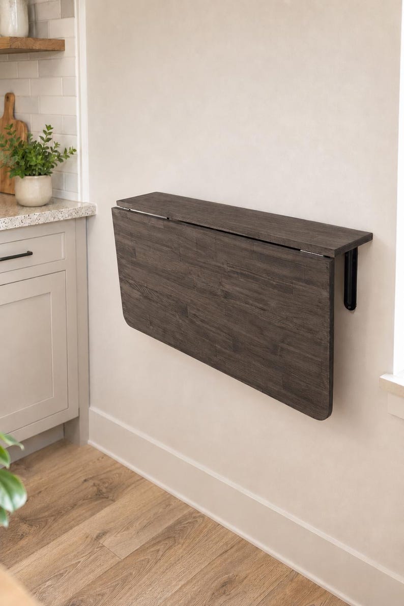 May include: A wall-mounted, dark gray wood-grain drop-leaf table featuring a rectangular top and a hinged panel that folds downward. The table is secured to the wall with black metal brackets, providing a space-saving surface for various uses.