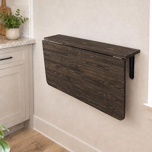 Op de afbeelding: Een aan de muur gemonteerde klaptafel met een donkerbruine houtnerfafwerking. Het rechthoekige oppervlak is voorzien van scharnieren om naar beneden tegen de muur te klappen, ondersteund door een zwart metalen beugelmechanisme aan de zijkant.