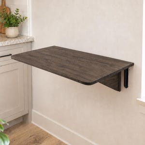 May include: A wall-mounted, rectangular, dark wood-toned folding table featuring rounded corners and a smooth surface. The table is attached to the wall with black metal support brackets, allowing it to be positioned horizontally for use.