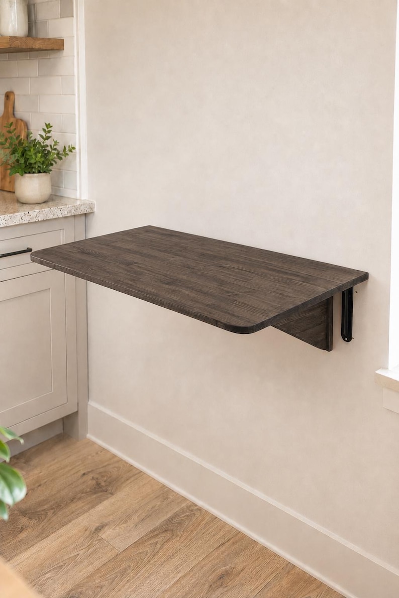 May include: A wall-mounted, rectangular, dark wood-toned folding table featuring rounded corners and a smooth surface. The table is attached to the wall with black metal support brackets, allowing it to be positioned horizontally for use.