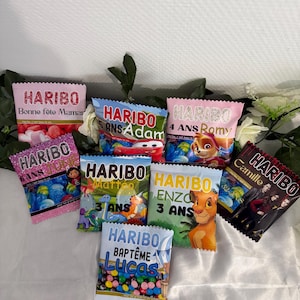 Peut inclure: Un ensemble de sept petits sachets de bonbons Haribo rectangulaires personnalisés avec des prénoms et des graphismes de personnages comme Cars, Pat' Patrouille et Le Roi Lion, incluant les textes Bonne fête Maman, 5 ANS Adam, 4 ANS Romy, 6 ANS June, 3 ANS Matteo, 3 ANS Enzo, Camille et BAPTÊME Lucas.