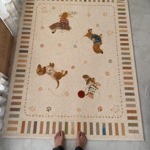 Puede incluir: Una alfombra rectangular con base de color crema que presenta un borde de rayas verticales alternas en tonos tostado, marr&oacute;n y azul. El centro muestra cuatro perros salchicha ilustrados en varias poses, acentuados por peque&ntilde;as huellas, estrellas y una pelota roja.