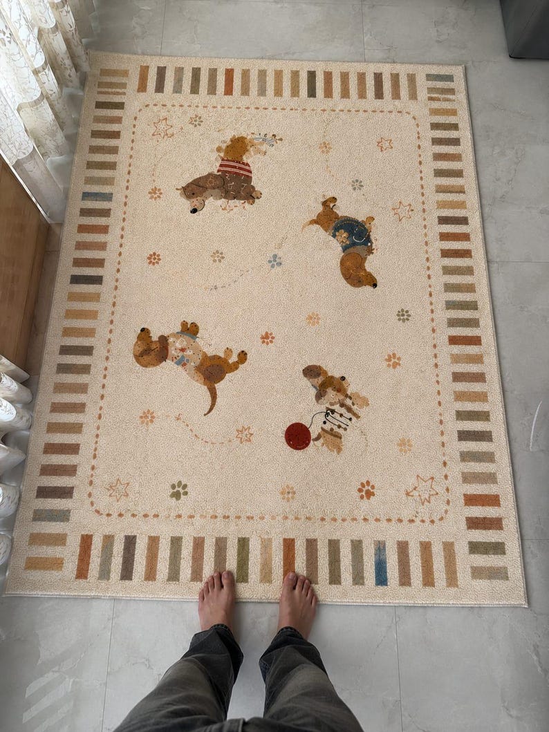 Puede incluir: Una alfombra rectangular con base de color crema que presenta un borde de rayas verticales alternas en tonos tostado, marr&oacute;n y azul. El centro muestra cuatro perros salchicha ilustrados en varias poses, acentuados por peque&ntilde;as huellas, estrellas y una pelota roja.