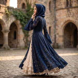 Könnte beinhalten: Ein bodenlanges Kleid mit einem dunkelblauen Spitzenüberwurf, einer Kapuze und langen, ausgestellten Ärmeln, getragen über einem cremefarbenen Rock mit mehreren Rüschenschichten. Der Spitzenüberwurf ist vorne offen.