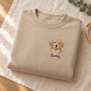 May include: A beige crewneck sweatshirt featuring a detailed embroidered portrait of a golden retriever on the left chest. Below the dog's face, the name Bailey is stitched in a dark maroon cursive font, creating a personalised design.