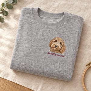May include: A heather grey crewneck sweatshirt featuring an embroidered illustration of a golden-brown curly-haired dog on the chest, with the text "doodle mama" stitched in dark red thread directly below the dog's face.