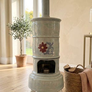 May include: A light blue ceramic stove with a floral design. The stove has a cylindrical body with a decorative flower and scrollwork. It stands on ornate silver-colored feet. A small olive tree in a terracotta pot is in the background.