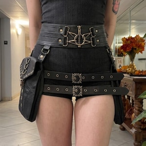 May include: A black utility belt featuring a wide waistband with a central zip and metallic hardware, two attached side pouches, and two horizontal leg straps with ornate buckles. The belt is worn over a black ribbed top and matching shorts.