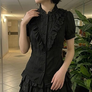 Puede incluir: Una blusa negra de manga corta con cuello alto de volantes, una fila vertical de pequeños botones negros en el centro y un adorno de volantes decorativo que se extiende desde los hombros hasta la cintura. La tela tiene un patrón texturizado.