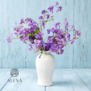 May include: A white ceramic vase with vertical fluted ridges containing a bouquet of vibrant purple flowers with four petals each. The vase features a black geometric logo and the text ALEXA printed in black on the lower left side of the frame.