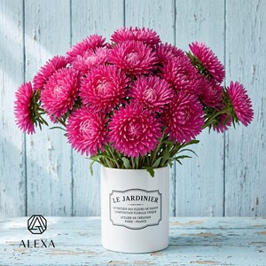 May include: A bouquet of vibrant magenta aster flowers arranged in a white cylindrical ceramic vase. The vase features a black label with the text LE JARDINIER, ENTRETIEN DES FLEURS DE SAISON, COMPOSITION FLORALE UNIQUE, ATELIER DE CRÉATION, PARIS - FRANCE.