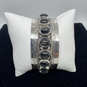 May include: A wide silver cuff bracelet featuring a central vertical row of seven smooth, oval-shaped black stones. The silver band is decorated with intricate stamped sunburst patterns, small silver beads, and twisted wire detailing along the edges.