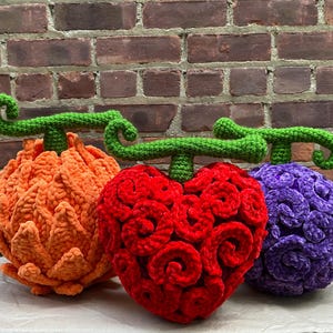 May include: Three handmade crochet plushies shaped like stylized fruits. The left is orange with pointed textures, the center is a heart-shaped red fruit with swirling patterns, and the right is a round purple fruit with a textured, curly surface.