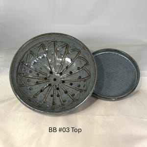 Può includere: Una ciotola per frutti di bosco in ceramica blu-grigio con base perforata e un piattino circolare piatto abbinato. L'interno della ciotola presenta un motivo radiale di linee incise e fori di drenaggio. Il testo BB #03 Top è stampato sotto.