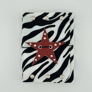 May include: A rectangular journal cover featuring a black and white zebra print pattern. The cover is accented with silver-toned metal studs along the top and bottom edges and a central dark red star-shaped closure with silver studs.