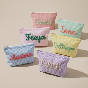 Puede incluir: Seis estuches rectangulares con cremallera hechos de tela seersucker en varios colores pastel, cada uno con un nombre bordado en un color contrastante. Los estuches llevan los nombres Celeste, Ione, Freya, Calliope, Aurora y Lilith.