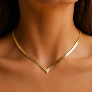 Puede incluir: Un collar de cadena de espiga en tono dorado con un diseño en forma de V. Los eslabones metálicos planos y flexibles crean una superficie lisa y reflectante que descansa sobre el cuello, formando una punta marcada en el centro de la clavícula.