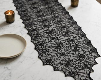 May include: A black lace table runner featuring an intricate, repeating spider web pattern with scalloped edges. The runner is elongated and rectangular, showcasing a delicate, semi-sheer mesh construction that displays a detailed web design.