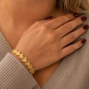 May include: A gold-toned bracelet featuring a series of connected four-leaf clover shapes. The metal surface has a textured, matte finish. The bracelet is worn around the wrist, showcasing a repeating floral-inspired link design in a bright yellow gold color.