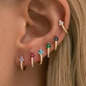 Puede incluir: Un conjunto de cinco pendientes de aro dorados, cada uno con un pequeño grupo de tres piedras preciosas. Los pendientes muestran una variedad de colores que incluyen morado claro, rosa oscuro, turquesa, verde y azul oscuro.