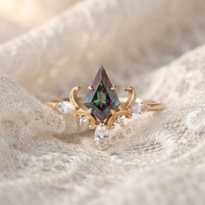 Puede incluir: Un anillo de oro con una piedra preciosa central en forma de cometa, multicolor con tonos verdes y morados iridiscentes. El engaste incluye detalles de filigrana dorada y está acentuado por cuatro pequeñas piedras claras de talla marquesa.