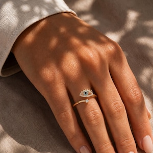 Puede incluir: Un anillo abierto de oro con un motivo en forma de ojo adornado con pequeñas piedras claras y una piedra azul oscuro en el centro, complementado con una piedra clara única en el extremo opuesto de la banda, creando un aspecto detallado.