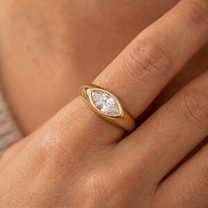Puede incluir: Un anillo de oro con una piedra preciosa transparente de talla marquesa engastada en un bisel liso y pulido. La banda es gruesa y redondeada, estrechándose ligeramente hacia los lados, con un acabado brillante que refleja la luz.