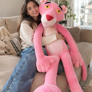 Puede incluir: Un peluche grande de la Pantera Rosa con pelaje rosa brillante, vientre de color rosa más claro, nariz roja y ojos amarillos con pupilas negras. El personaje tiene extremidades largas, una cola larga y orejas prominentes, diseñado como una figura suave.